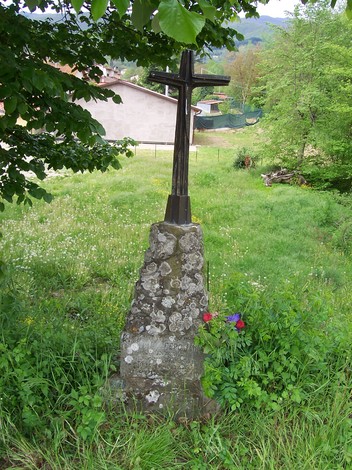Cippo commemorativo con croce, immerso nella natura, circondato da fiori e erba.