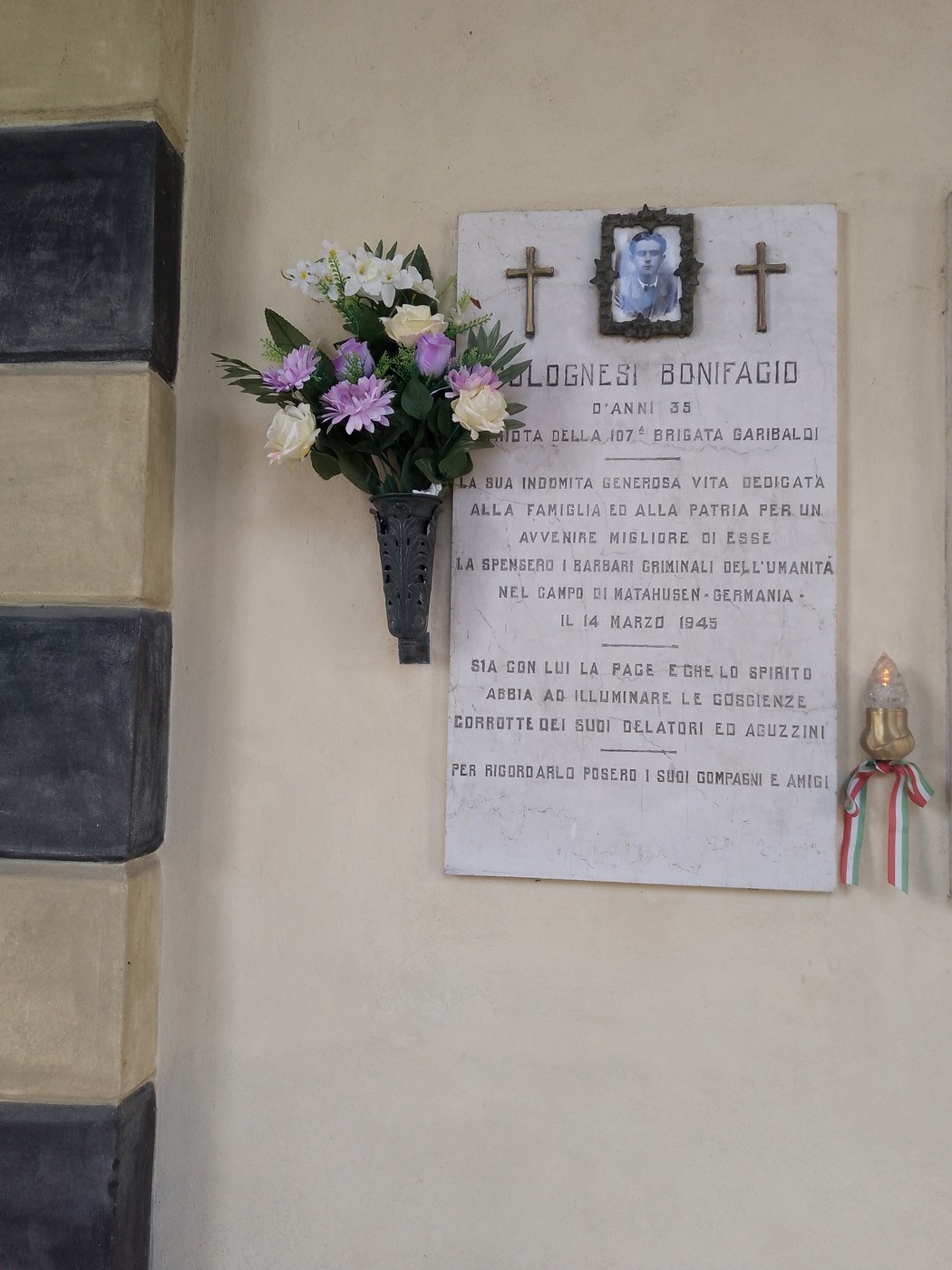 Lapide commemorativa a Colognesi con fiori e foto, in memoria di Bonifacio. Riflessione sulla vita e l'eredità.