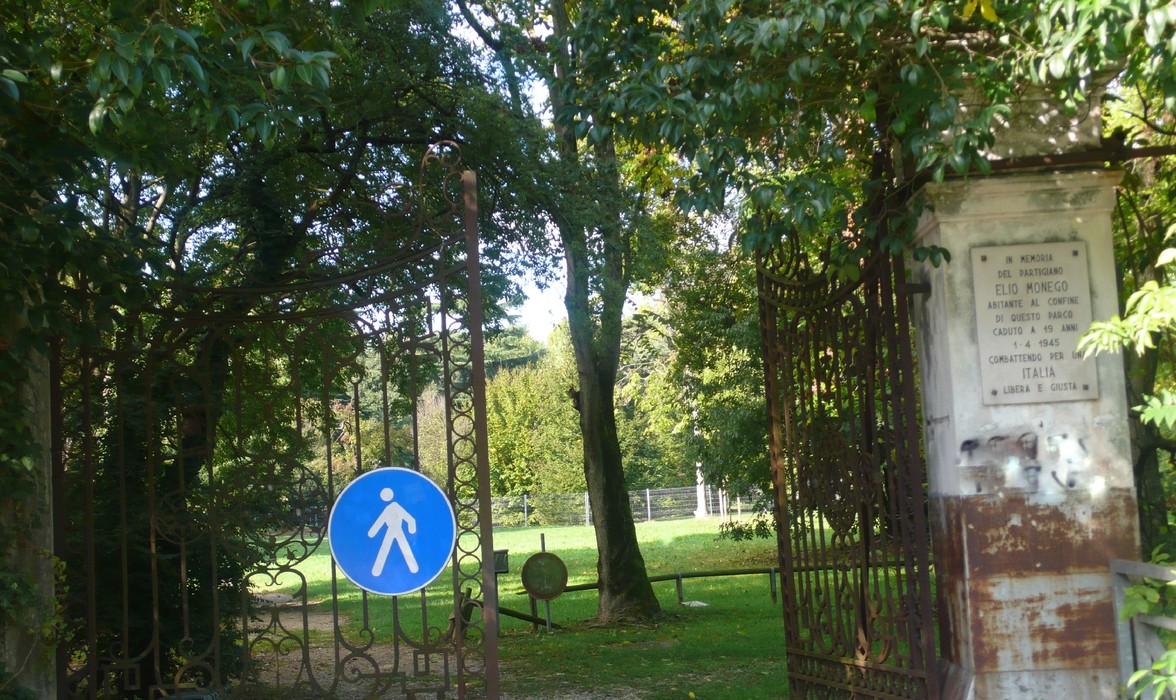 Ingresso del parco con chiusura in ferro e segnale di attraversamento pedonale su sfondo verde di natura.