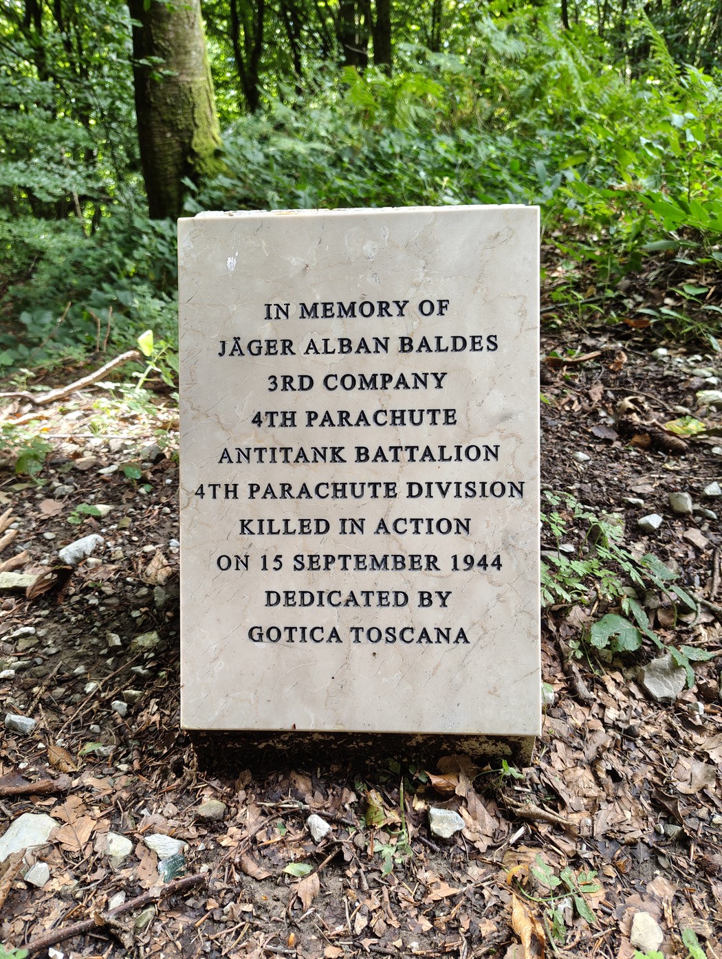 Cippo commemorativo dedicato a Jäger Alban Baldes, situato in un'area boschiva, ricco di vegetazione.