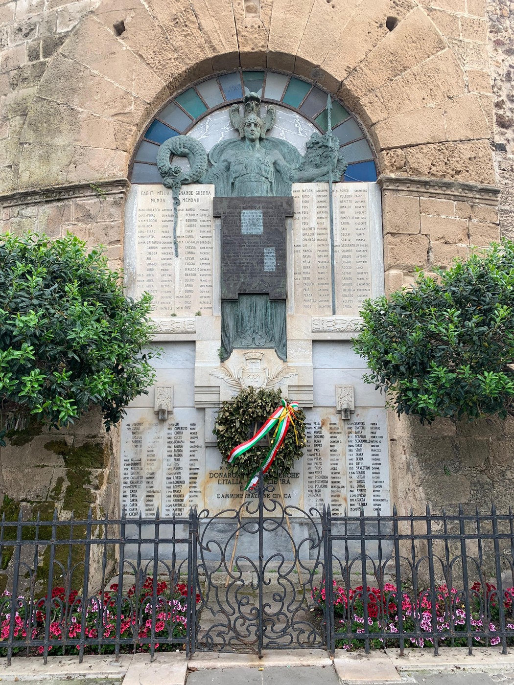 Monumento ai Caduti di Alghero, con statue e elenchi commemorativi, circondato da piante e fiori.