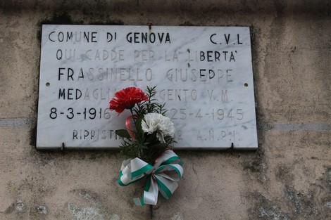 Lapide commemorativa dedicata a Giuseppe Frassinello, con fiori e nastri, affissa su un muro.