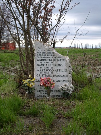 Cippo commemorativo dedicato alle vittime di un bombardamento, decorato con fiori e circondato da vegetazione.