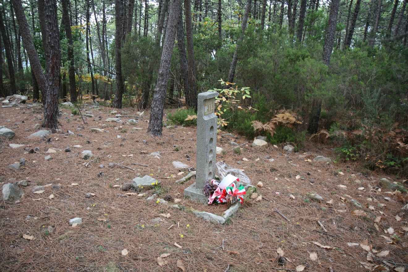Cippo commemorativo immerso nel bosco, circondato da alberi e foglie, con decorazioni floreali ai suoi piedi.