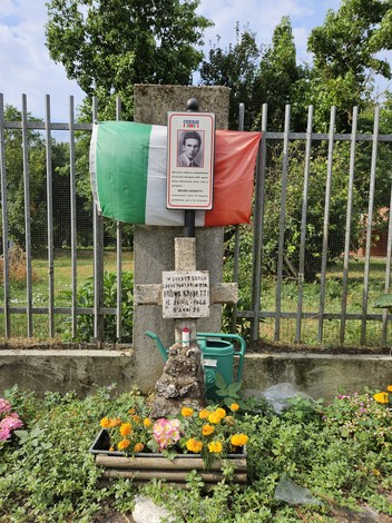 Croce commemorativa con foto e bandiera italiana, circondata da fiori e verde, in un contesto urbano.