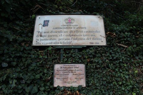 Targa commemorativa dedicata ai bambini vittime di una bomba, con iscrizioni e simboli religiosi, immersa nella vegetazione.