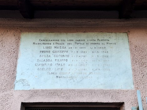 Lapide in marmo con lunga iscrizione e elenco di nomi, affissa su una parete esterna intonacata.
