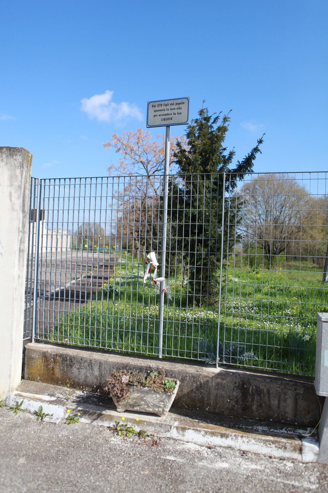 Targa commemorativa ai caduti del Poligono di tiro, situata all'ingresso di un'area verde recintata.