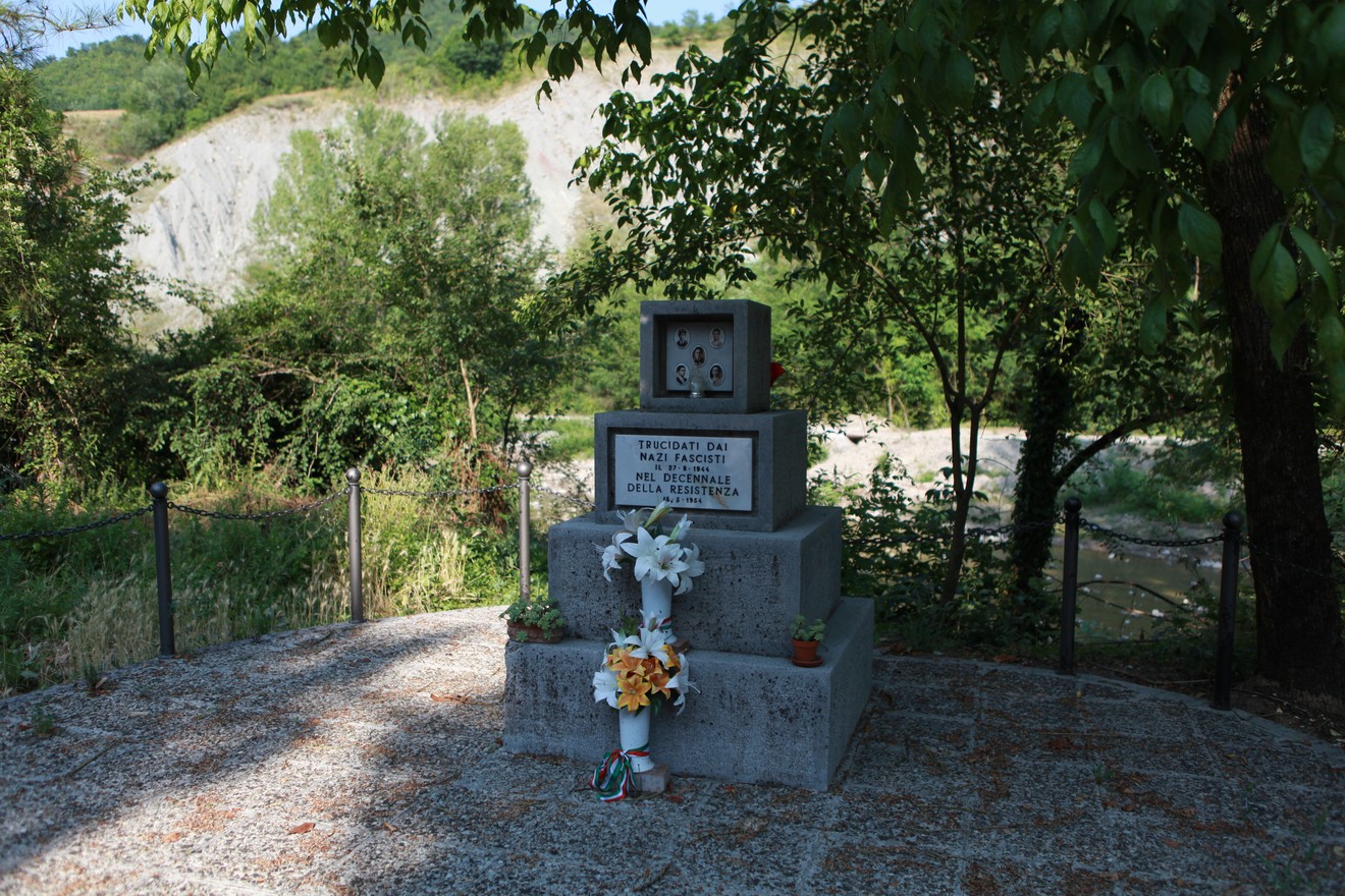 Monumento ai partigiani fucilati, circondato da vegetazione, con decorazioni floreali. Un luogo di memoria e rispetto.