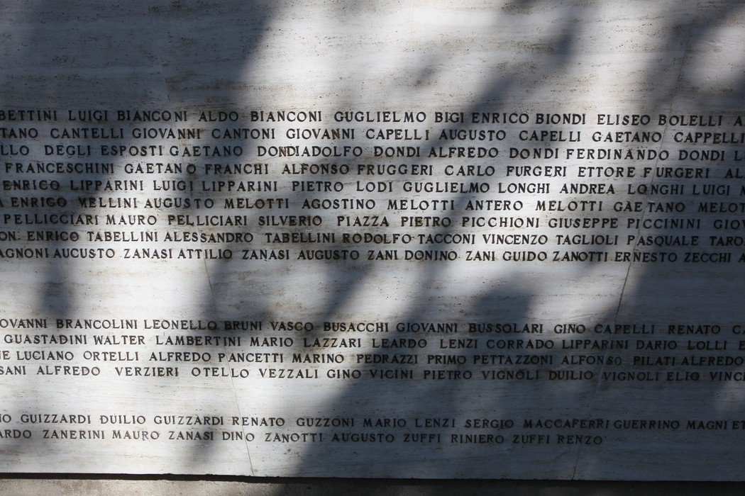 Monumento ai Caduti con iscrizioni commemorative su una superficie in pietra. Ombre delicate sovrastano il memoriale.