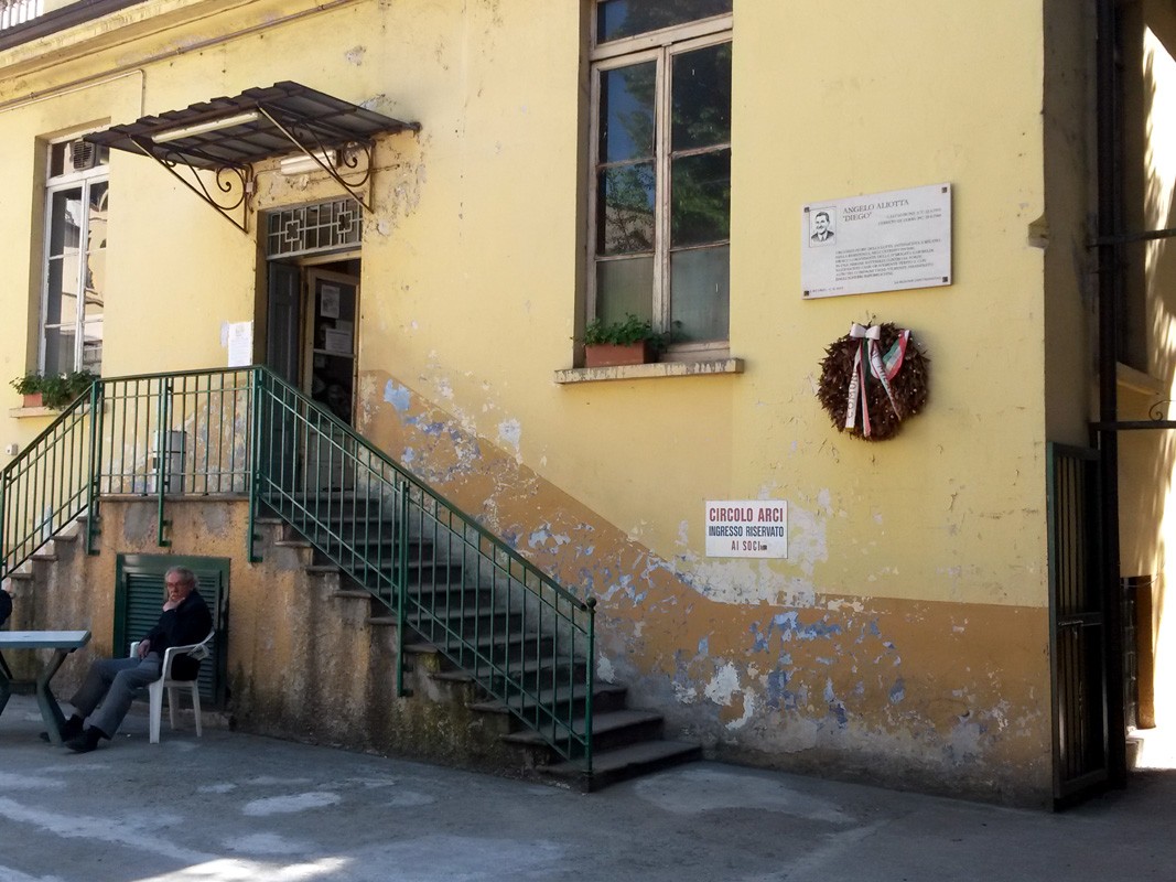 Lapide commemorativa ad Aliotta su un edificio giallo, decorata con una corona e circondata da un'atmosfera storica.