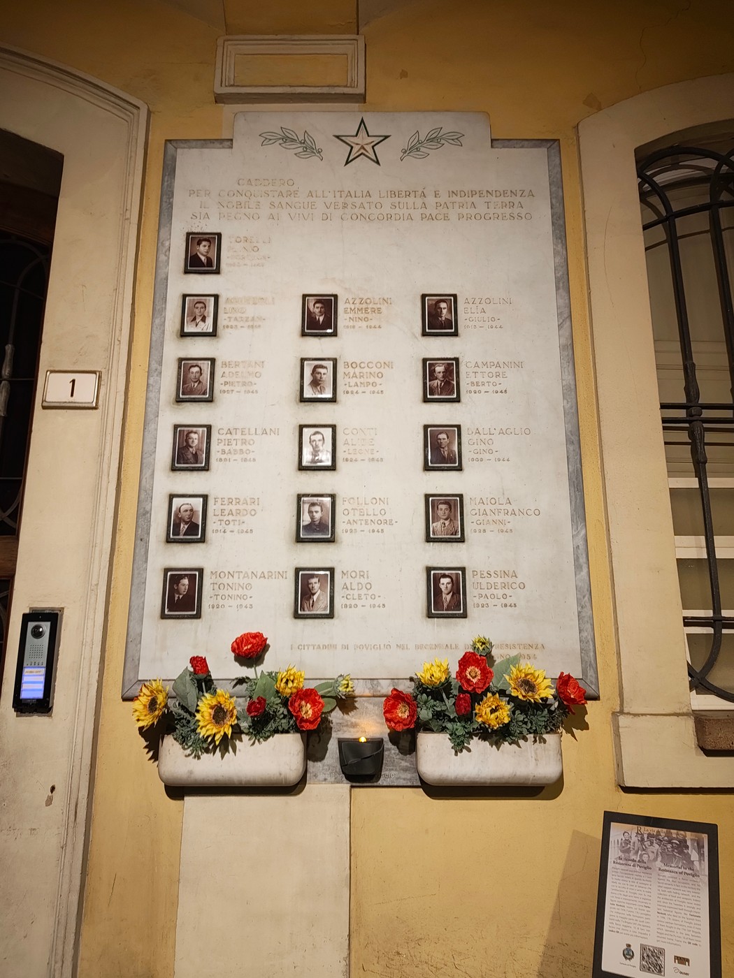 Lapide marmorea con foto e nomi dei caduti, stella in cima, fiori e lumino sottostanti.