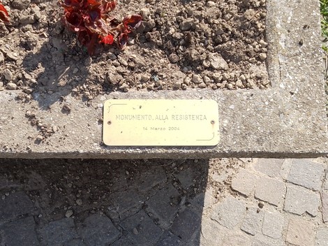 Monumento alla Resistenza con placca commemorativa, circondato da aiuole e pavimentazione in pietra.