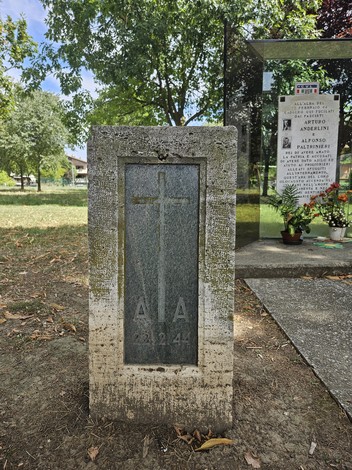Monumento commemorativo con iscrizioni e croce in evidenza, immerso in un'area verde.