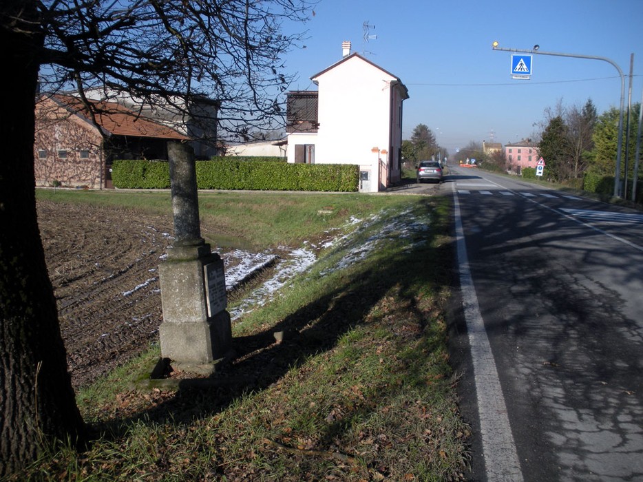 Cippo stradale situato lungo la strada, vicino a edifici residenziali e un campo.