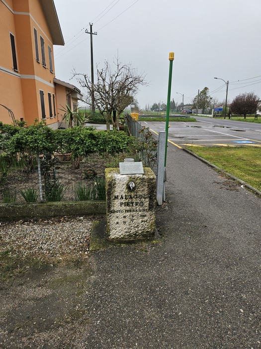 Cippo storico a Malagoli, accanto a una strada e vegetazione, con un contesto urbano in lontananza.