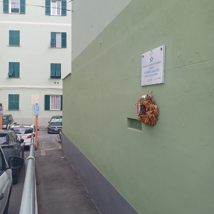 Lapide con iscrizione su parete verde, ornata da una corona secca appesa sotto il bordo.