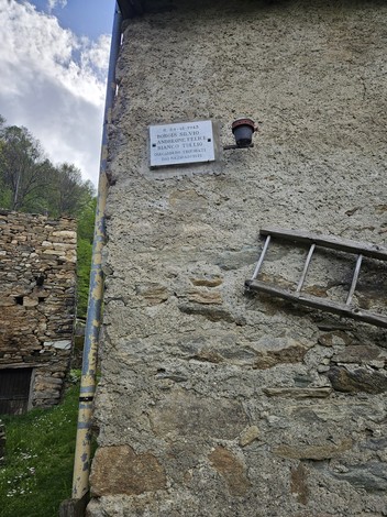 Lapide commemorativa su un muro di pietra, con elementi architettonici rustici nei dintorni.