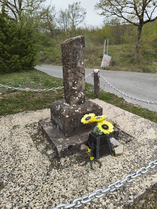 Cippo memoriale in pietra circondato da fiori, situato in un'area naturale con strada asfaltata sullo sfondo.