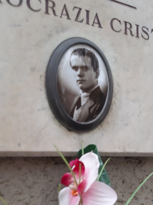 Lapide commemorativa con fotografia in bianco e nero di un giovane, decorata da un fiore in primo piano.
