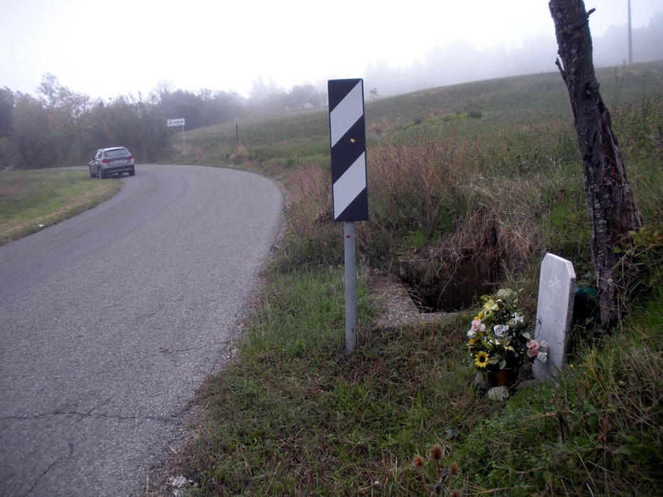 Cippo commemorativo ai Rufinotti vicino a una curva stradale, circondato da fiori e vegetazione.