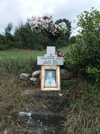 Una croce commemorativa con fiori artificiali e un'immagine incorniciata, situata in un'area verde.