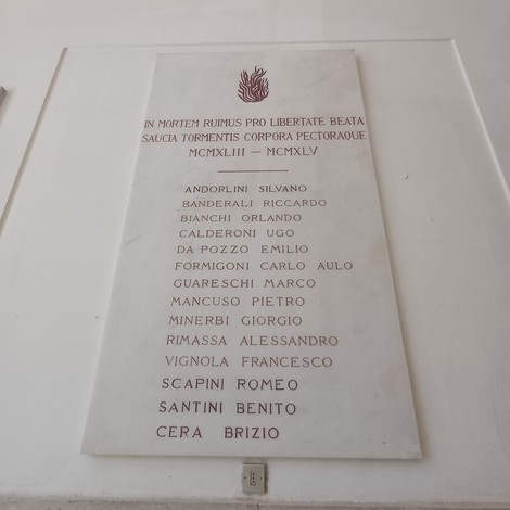 Lapide in marmo su parete bianca, con fiamma incisa e lungo elenco di nomi dei caduti.
