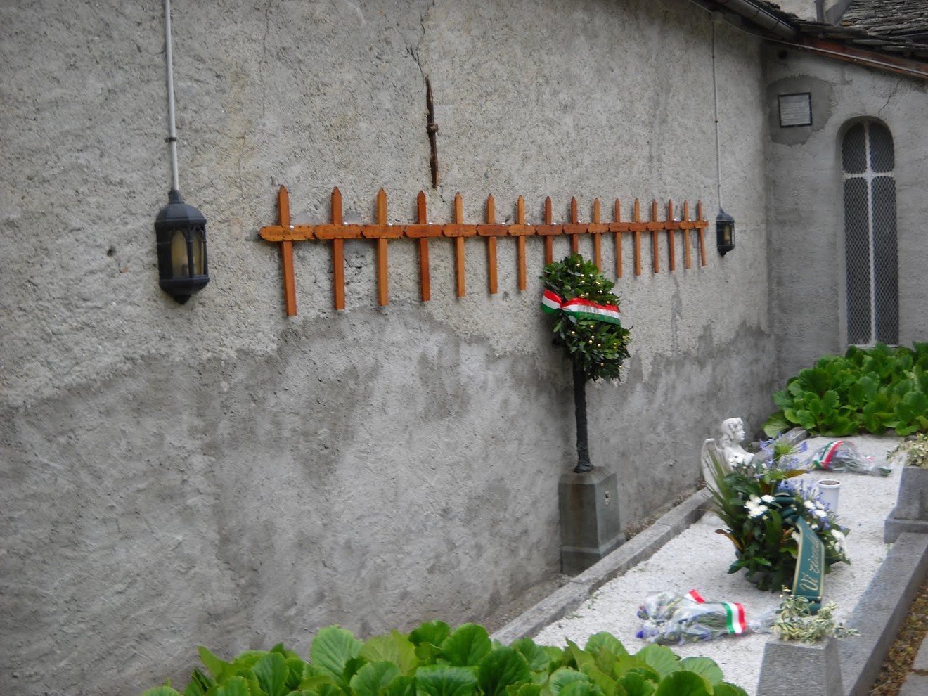 Sacrario dei Caduti nel cimitero, con decorazioni e omaggi floreali su un muro semplice.