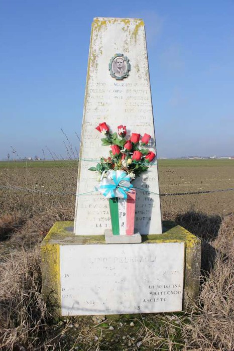 Cippo commemorativo a Pederzoli, ornato con fiori rosso e verde, immerso in un paesaggio rurale.