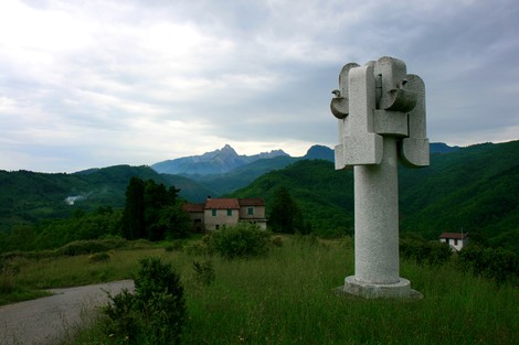 Monumento di Valla in un paesaggio collinare con montagne sullo sfondo e una casa immersa nella vegetazione.