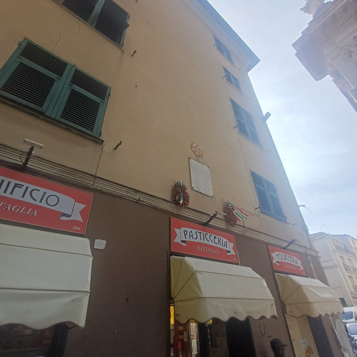 Facciata di un palazzo con lapide e iscrizione tra due finestre, sopra le insegne di una pasticceria.