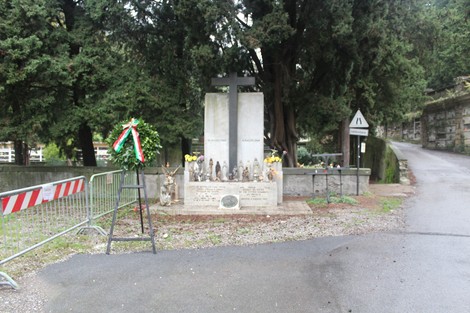 Monumento alle vittime civili, con croce e decorazioni floreali, circondato da alberi in un'area tranquilla.