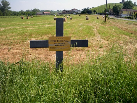Croce commemorativa di Ettore Minotti in un campo, circondata da un paesaggio rurale.