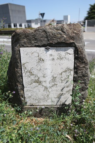 Cippo commemorativo in pietra dedicato alla strage di Pratomaggiore, circondato da vegetazione e un paesaggio urbano.