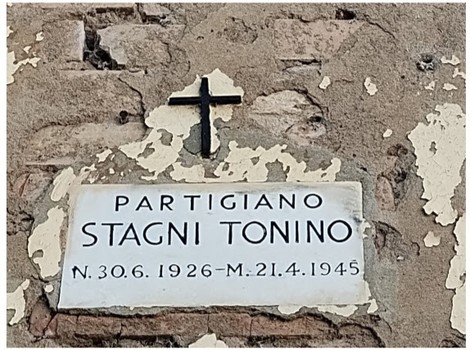 Lapide commemorativa di un partigiano, con croce, su una parete di mattoni in pietra.