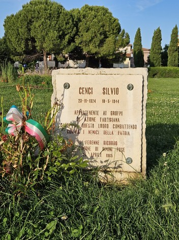 Cippo commemorativo dedicato a Silvio Cenci, situato in un'area verde, circondato da piante e decorazioni tricolori.
