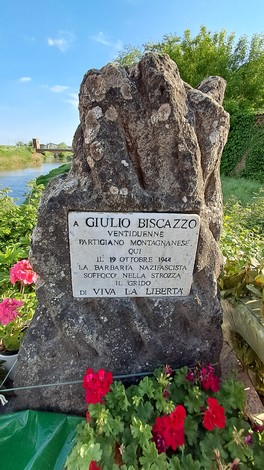 Cippo commemorativo dedicato a Giulio Biscazzo, con fiori colorati sullo sfondo in un ambiente naturale.