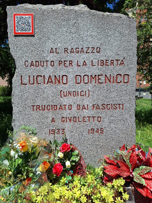 Cippo commemorativo in pietra dedicato a Luciano Domenico, caduto per la libertà, adornato di fiori.
