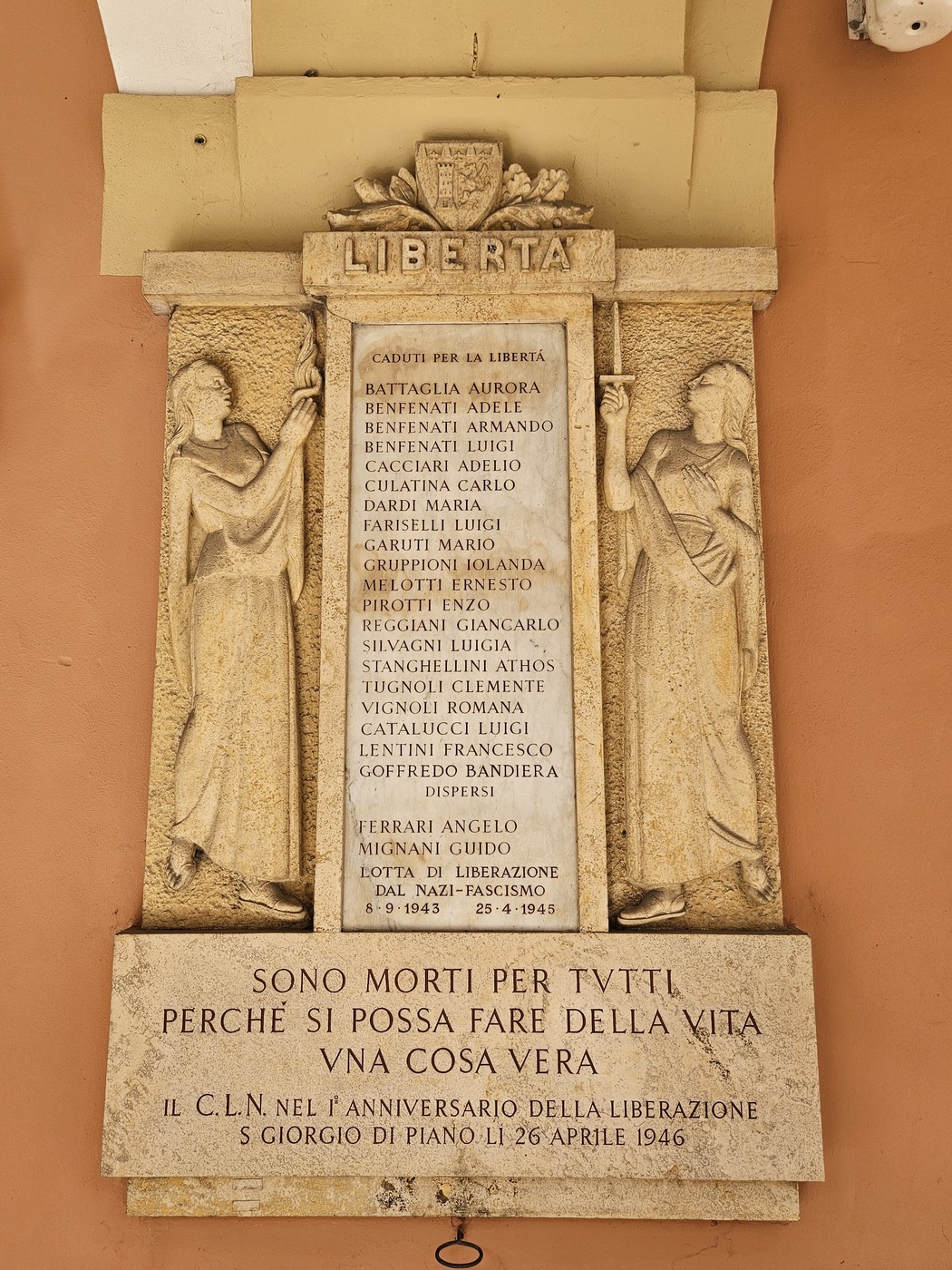 Lapide commemorativa con iscrizioni in onore dei caduti per la libertà, adornata da figure simboliche.