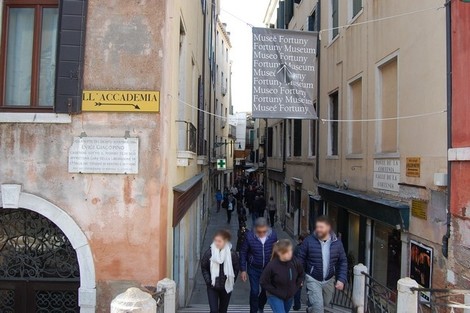 Vista di una calle di Venezia con visitatori che si avviano verso il Museo Fortuny, tra edifici storici.