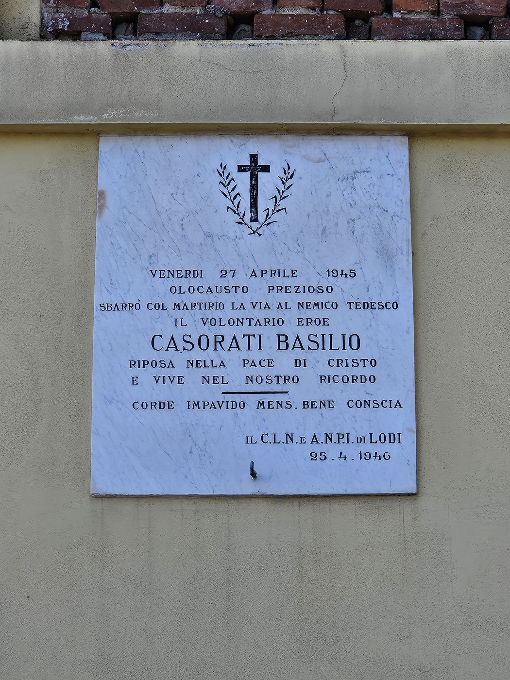Lapide marmorea con croce e corona d'alloro incisa, fissata su parete esterna, con iscrizione dedicatoria.