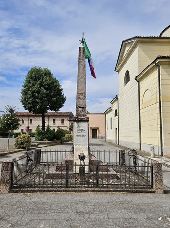 Monumento ai Caduti di Spineda, un obelisco con bandiere, circondato da una recinzione in un'area aperta.