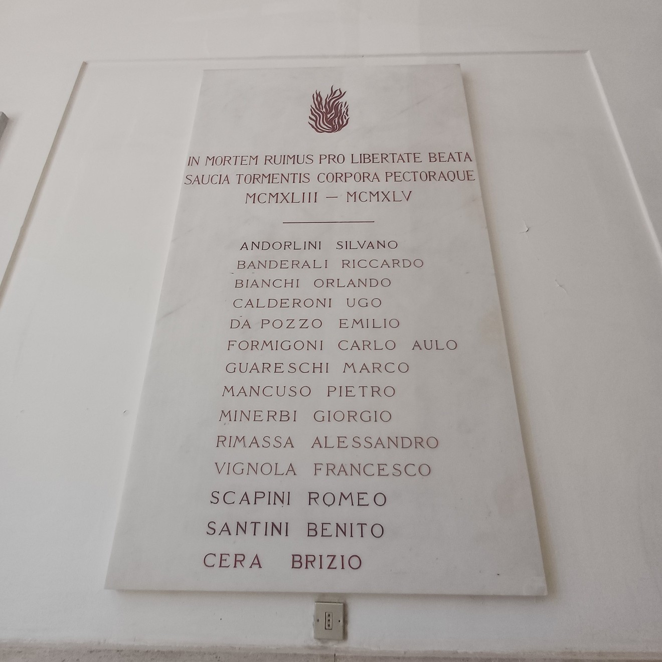 Lapide in marmo su parete bianca, con fiamma incisa e lungo elenco di nomi dei caduti.