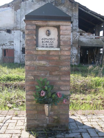 Cippo commemorativo dedicato ad Armando Bonezzi, con fiori e una foto, in un contesto di rovine.