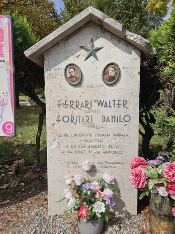 Cippo commemorativo con fotografie di Ferrari e Fornari, circondato da fiori e vegetazione.