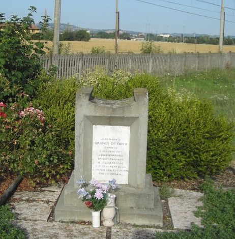 Cippo dedicato a Grandi Ottavio, circondato da vegetazione e campi, con fiori e candela al piede.