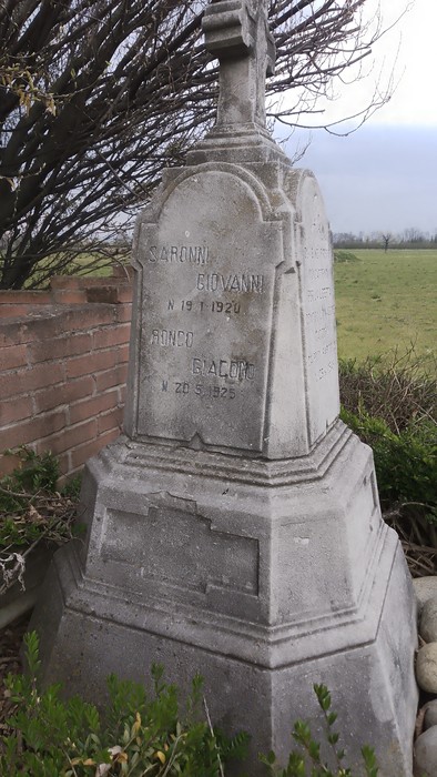 Cippo commemorativo dedicato ai partigiani di Camuzzago, tra vegetazione e un campo aperto sullo sfondo.