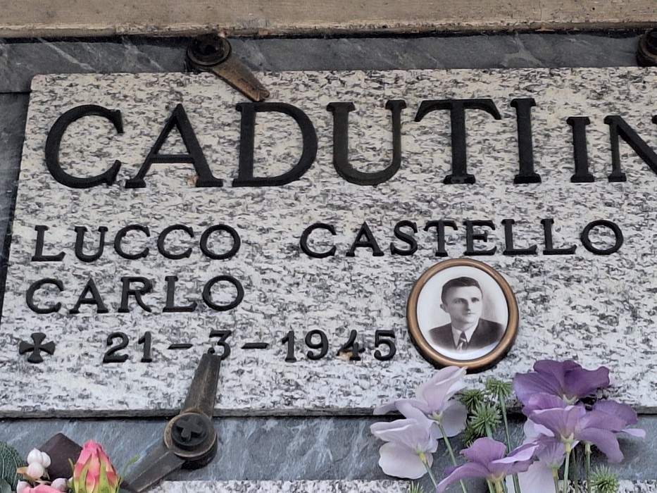 Lapide in marmo con iscrizione, ritratto fotografico ovale e fiori deposti sotto.