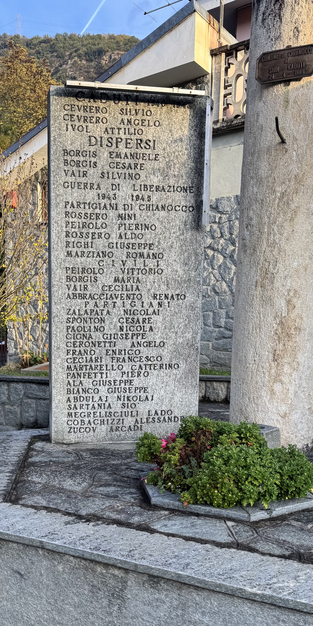 Stele in pietra con lunga iscrizione ai caduti, posta all'aperto davanti a un edificio, con cespuglio fiorito alla base.