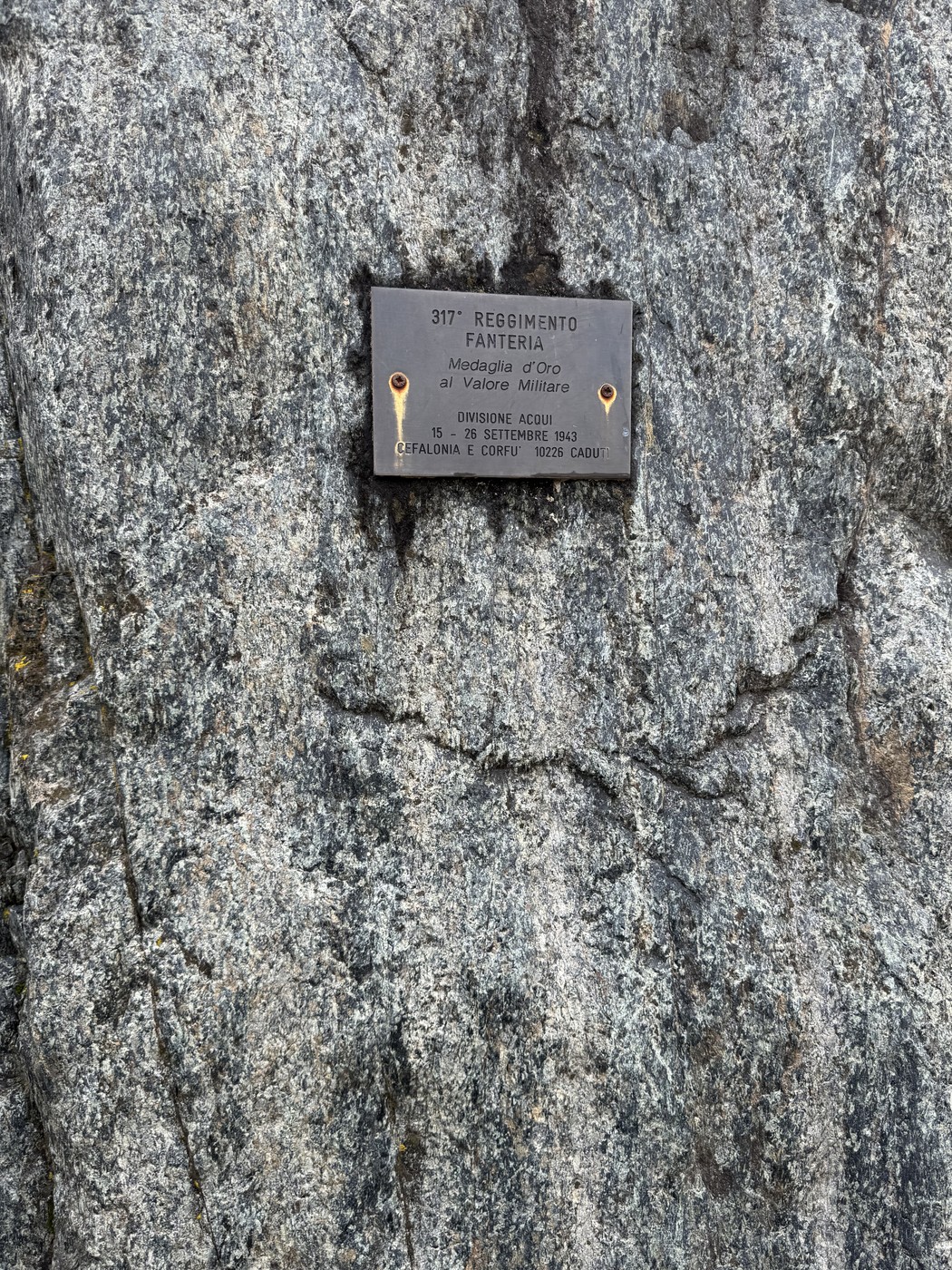 Targa metallica con iscrizione fissata su una parete di roccia grigia.